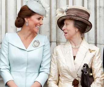 Two women in elegant attire and hats smiling at each other against a stone background.