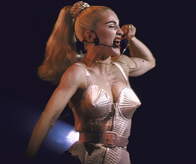 Madonna performing energetically on stage in a cone bra costume with a ponytail and headset microphone.