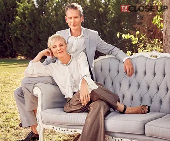 Couple seated on an elegant sofa outdoors, woman in white top, man in grey suit, serene garden setting.