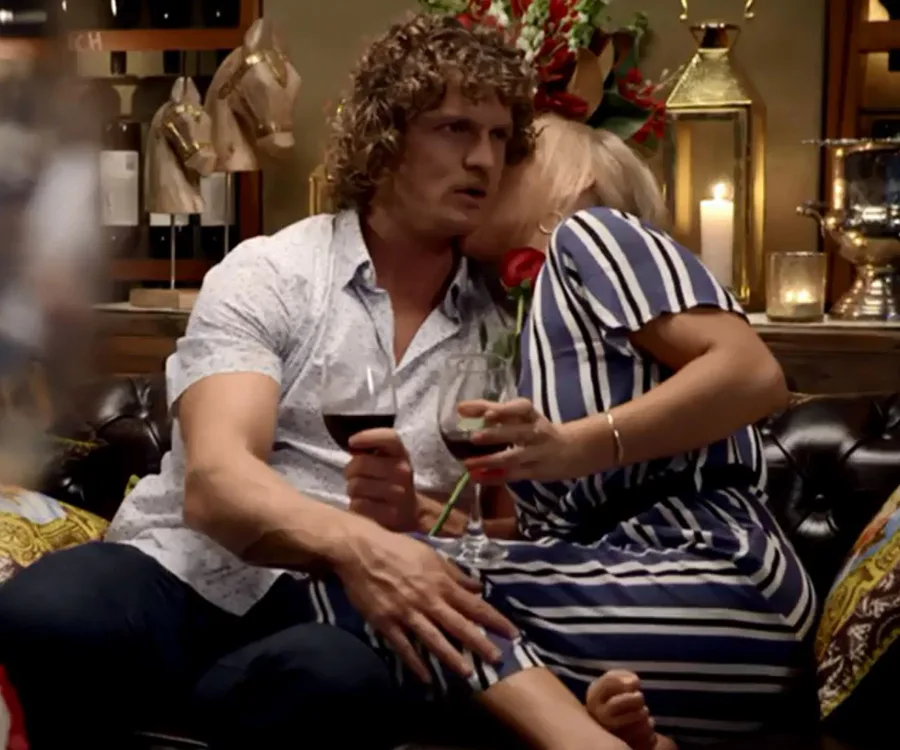 Man and woman, both holding wine glasses, sit closely on couch; woman leans in for kiss, rose in hand. Cozy interior.