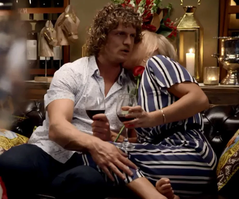 Man and woman, both holding wine glasses, sit closely on couch; woman leans in for kiss, rose in hand. Cozy interior.