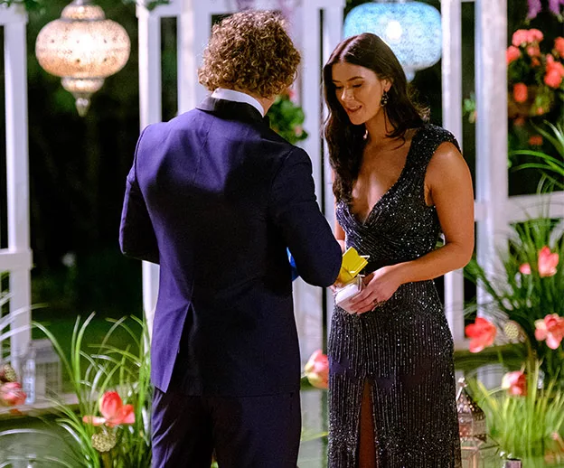 A man in a suit presents a yellow rose to a woman in a sparkling dress in a garden setting with lanterns and flowers.