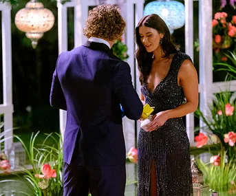 A man in a suit presents a yellow rose to a woman in a sparkling dress in a garden setting with lanterns and flowers.