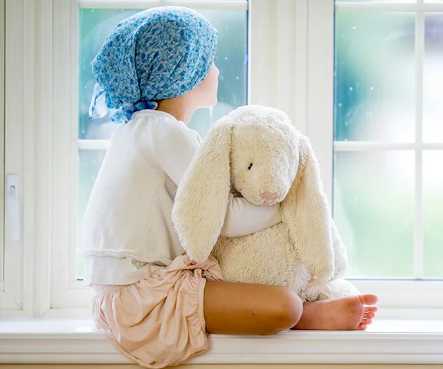 Child wearing headscarf holding plush toy, sitting by a window.
