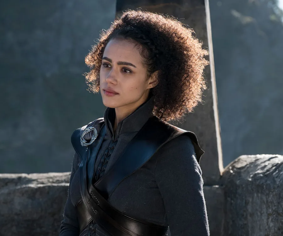 Missandei from "Game of Thrones" standing outdoors, wearing dark armor and looking thoughtful.
