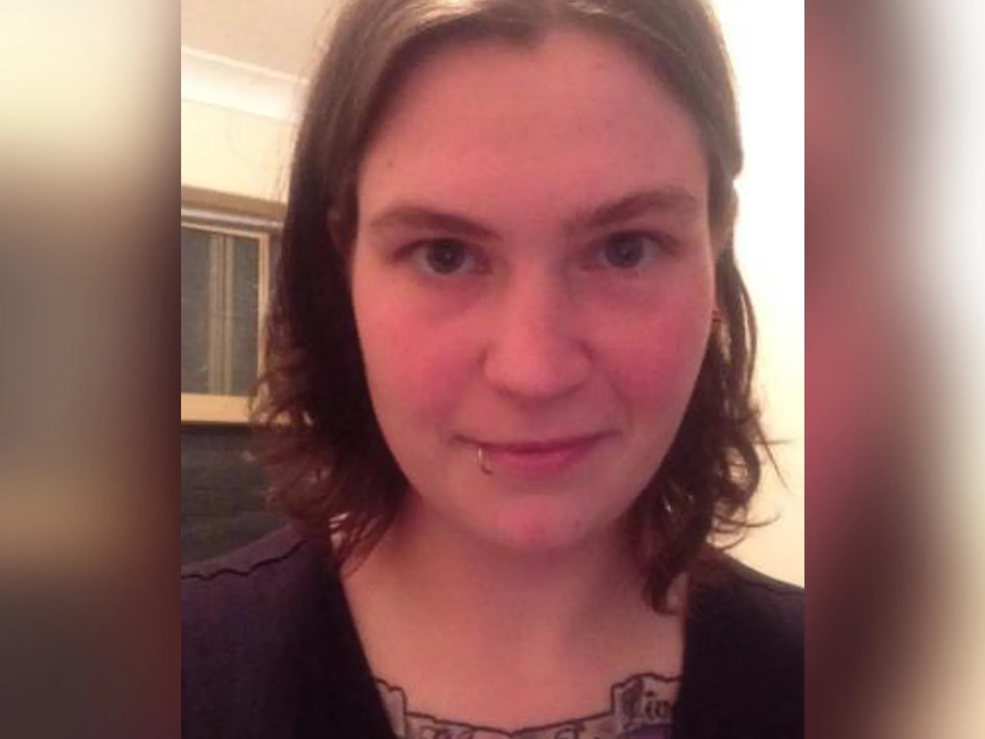 Woman with short hair, lip piercing, tattoo on chest, in indoor setting.