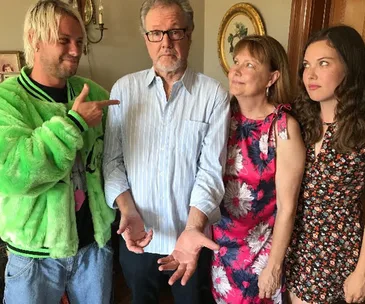 Man in green jacket points at serious older man with glasses, next to two smiling women in floral dresses.
