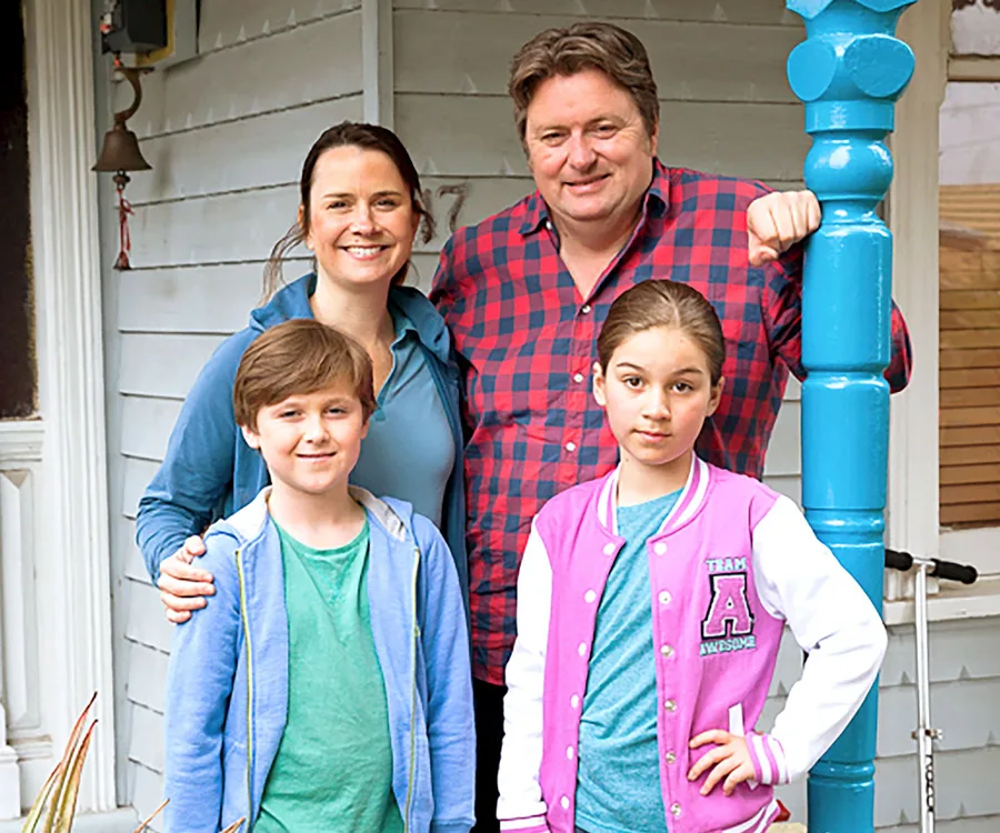 A family of four stands outside a house, smiling. The man wears a red plaid shirt; the others wear casual blue and pink clothing.