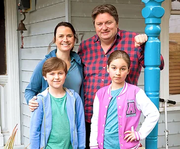 A family of four stands outside a house, smiling. The man wears a red plaid shirt; the others wear casual blue and pink clothing.