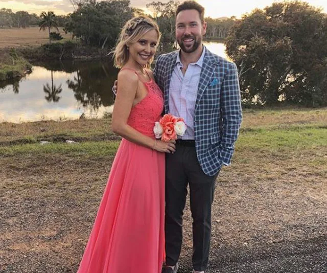 A couple poses by a scenic lake; the woman in a coral gown holding flowers, and the man in a plaid jacket smiles.