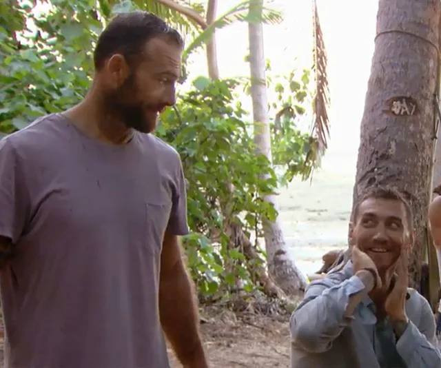 Two men in a tropical setting, one standing, the other crouched by a tree, both smiling and interacting.