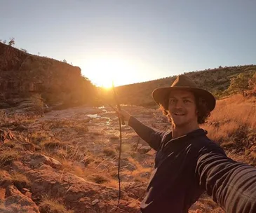 Selfie of a man in a hat holding a stick, in a rocky landscape at sunset.