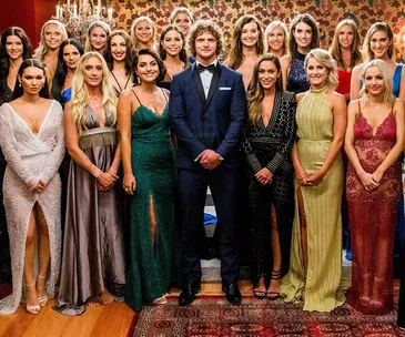 Contestants from "The Bachelor" gather in formal attire around the central male lead, posing together in a luxurious setting.