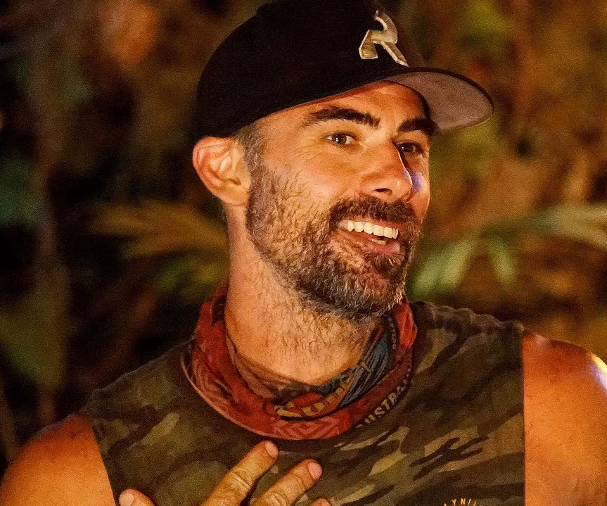 Man with a beard wearing a cap and camouflage shirt, smiling in a jungle setting.