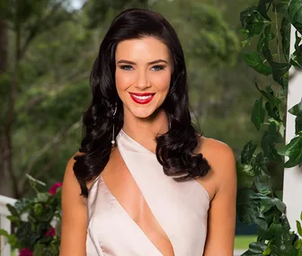 A woman with long dark hair and red lipstick smiles brightly outdoors, surrounded by greenery.