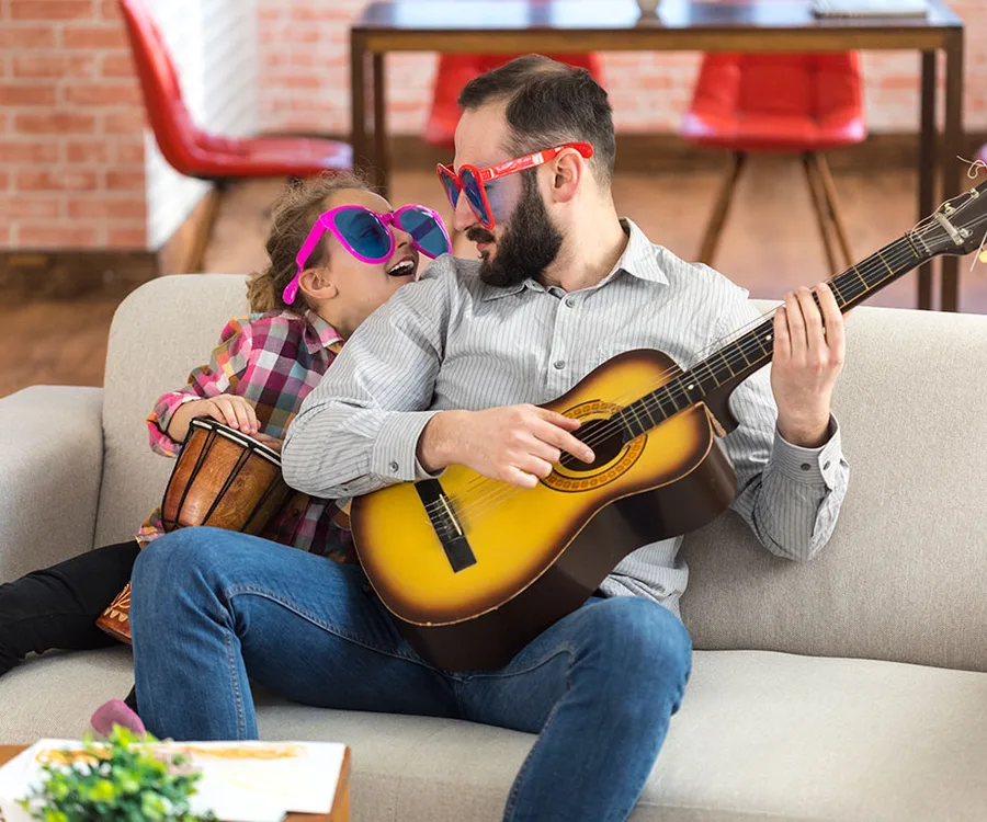 Man and child wearing sunglasses playing guitar and bongo on a couch, enjoying a musical moment together.