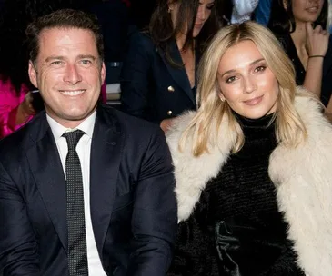 A man in a suit and a woman in a fur coat sit smiling at a fashion show audience.