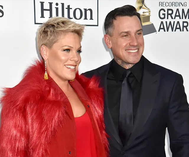Two people smiling at an event, one in a red outfit and the other in a dark suit, standing in front of a Grammy backdrop.