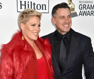 Two people smiling at an event, one in a red outfit and the other in a dark suit, standing in front of a Grammy backdrop.