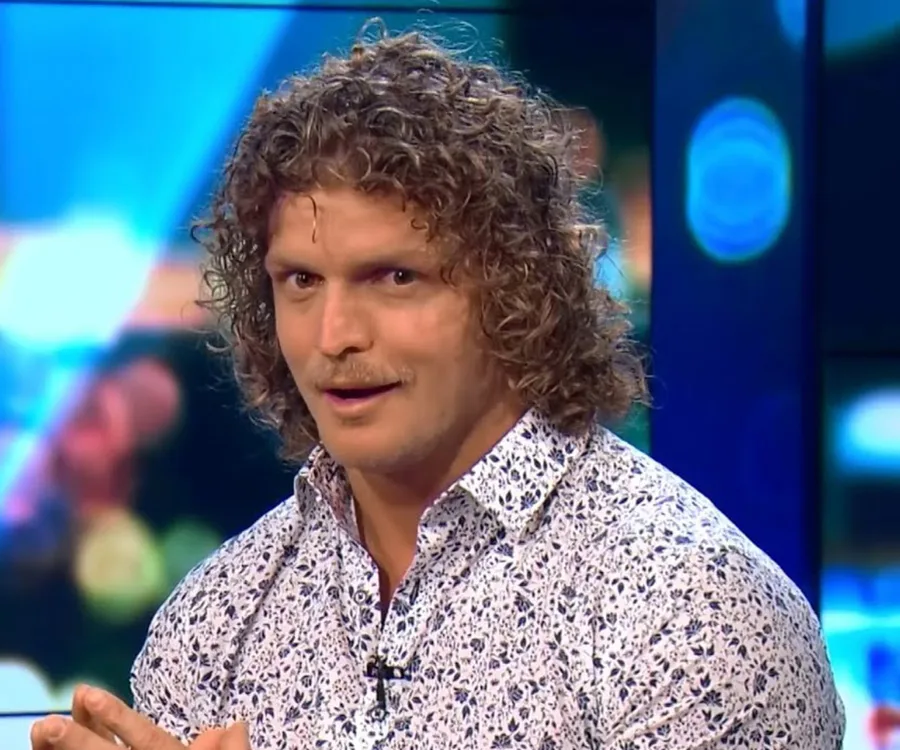 Man with curly hair and floral shirt looks at the camera with a surprised expression, colorful background behind.