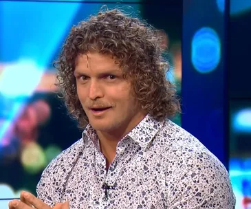 Man with curly hair and floral shirt looks at the camera with a surprised expression, colorful background behind.