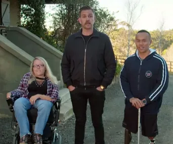Three people outdoors: a woman in a wheelchair, a man standing in the center, and another man with a prosthetic leg.