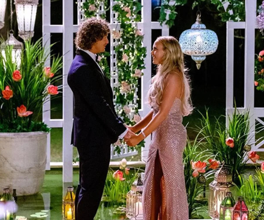 A couple holding hands in a romantic garden setting with lanterns and flowers at night.