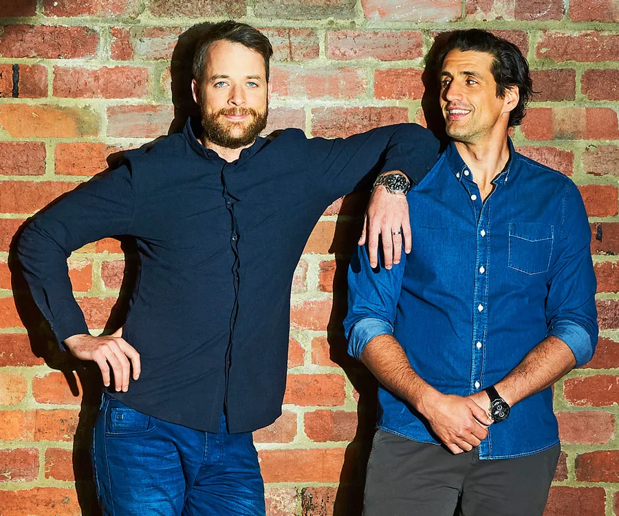 Two men smiling, standing against a brick wall. One man rests his arm on the other's shoulder.