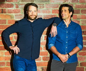 Two men smiling, standing against a brick wall. One man rests his arm on the other's shoulder.