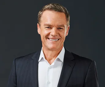 A smiling middle-aged man in a dark suit and white shirt against a dark background.