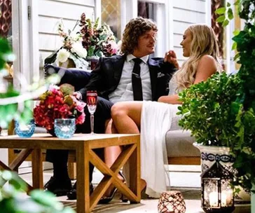 A man and woman sit closely on an outdoor sofa, surrounded by plants and flowers, with drinks on a wooden table.