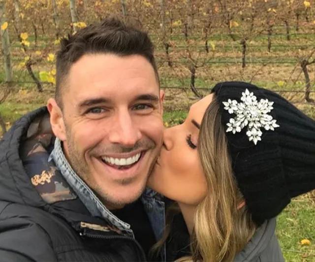 Man smiling as woman wearing a jeweled beanie kisses his cheek in a vineyard.