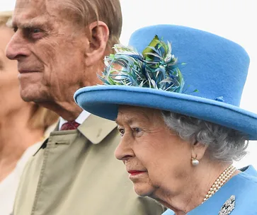 Senior couple at event, woman in ornate blue hat with feathers, man in tan overcoat, focused on the scene.