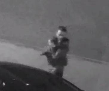 Black and white CCTV image shows a person holding a child near a parked vehicle.
