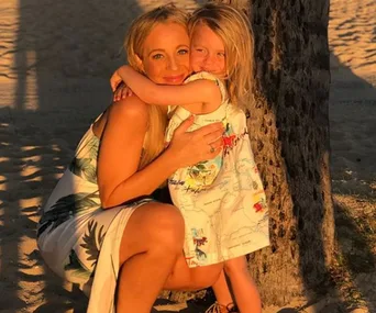 A woman and a young girl embrace by a tree on a sunlit beach, both smiling warmly.