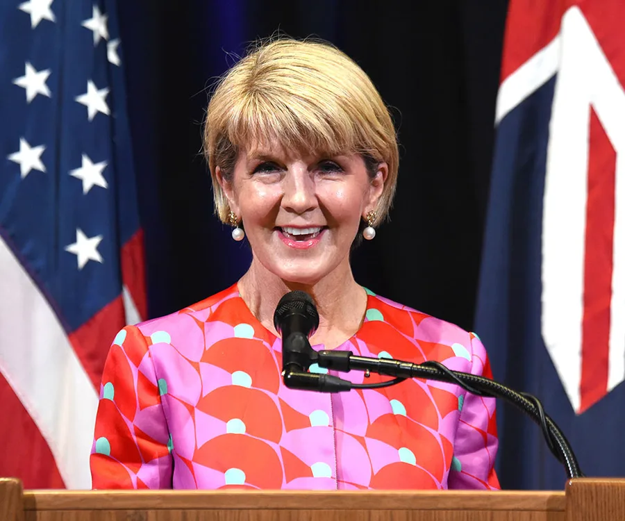 A woman speaks at a podium, with U.S. and Australian flags in the background, wearing a colorful patterned outfit.
