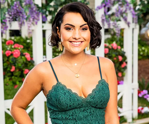 Young woman in a green lace dress smiles in a garden setting with flowers and white trellis in the background.