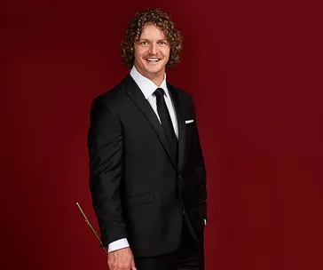 Man in a suit holding a rose, smiling against a red background.
