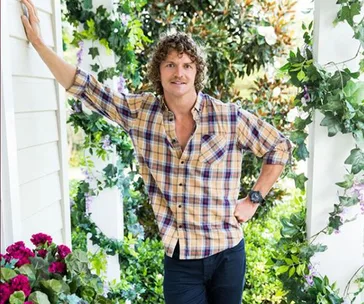 A person with curly hair in a plaid shirt stands in a garden setting, surrounded by greenery and flowers.