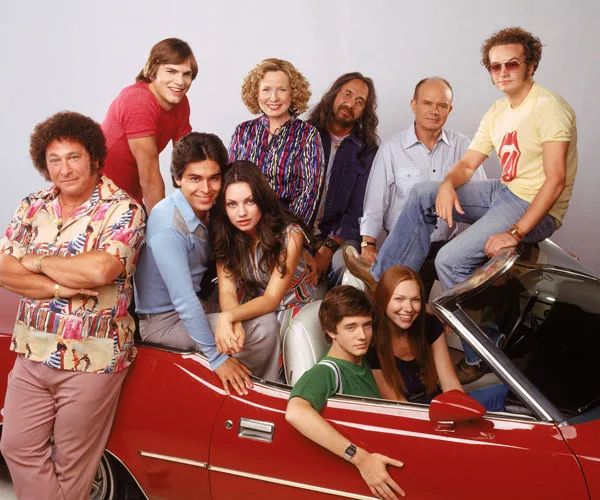 A group photo of "That '70s Show" cast posing around a red vintage car, smiling.
