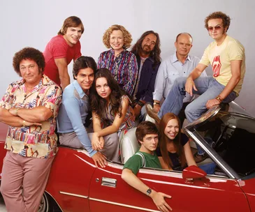 A group photo of "That '70s Show" cast posing around a red vintage car, smiling.