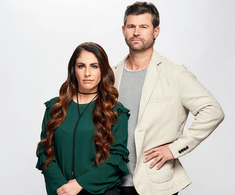 Two individuals, a woman in green and a man in a beige jacket, standing together against a plain background.