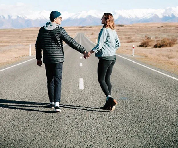 Couple holding hands walking down a road with snow-capped mountains in the background.