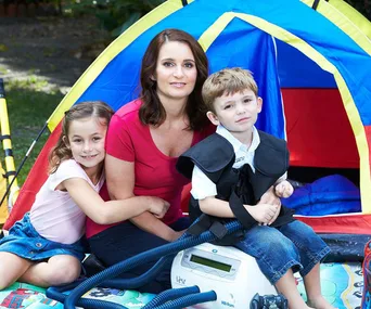 Mother with two children outside a colorful tent; one child uses medical equipment.