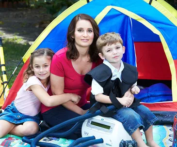 Mother with two children outside a colorful tent; one child uses medical equipment.