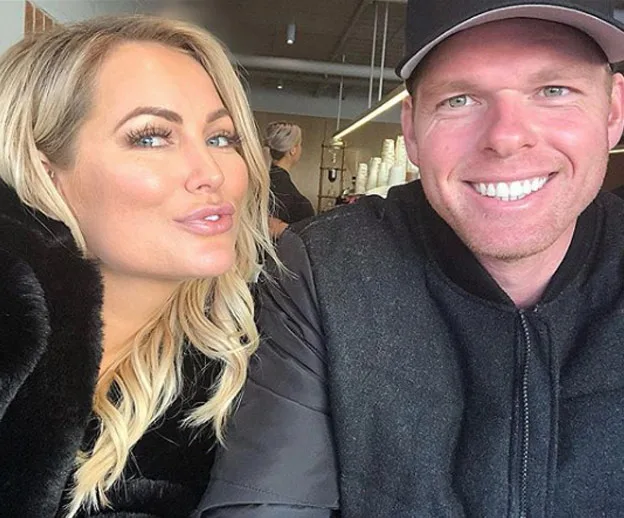 A woman with long blonde hair poses next to a smiling man in a cap inside a cafe.