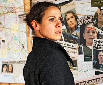 A woman in profile looks worried, standing before a wall covered with maps and newspaper clippings.
