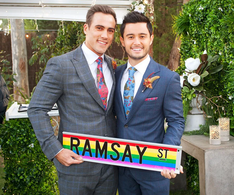 Two grooms smiling, dressed in suits, holding a "Ramsay St" rainbow sign, surrounded by greenery at a wedding.