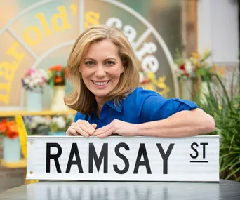Woman in a blue shirt smiling, posing with a "Ramsay St" sign in front of a café background.