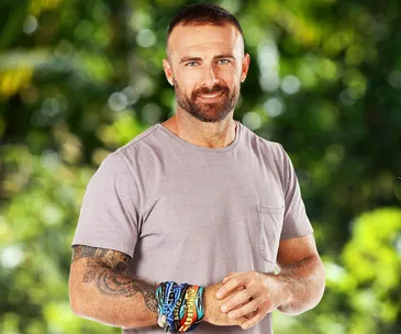Man with a beard wearing a pink shirt, sporting colorful wristbands, outdoor background with blurred greenery.